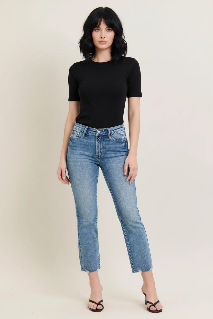 A person modeling a pair of light blue cropped denim jeans with a straight, slightly tapered leg and raw frayed hems, paired with black thong sandals against a plain beige studio background.