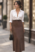Person wearing a white blouse and brown skirt standing in front of a building with large windows.