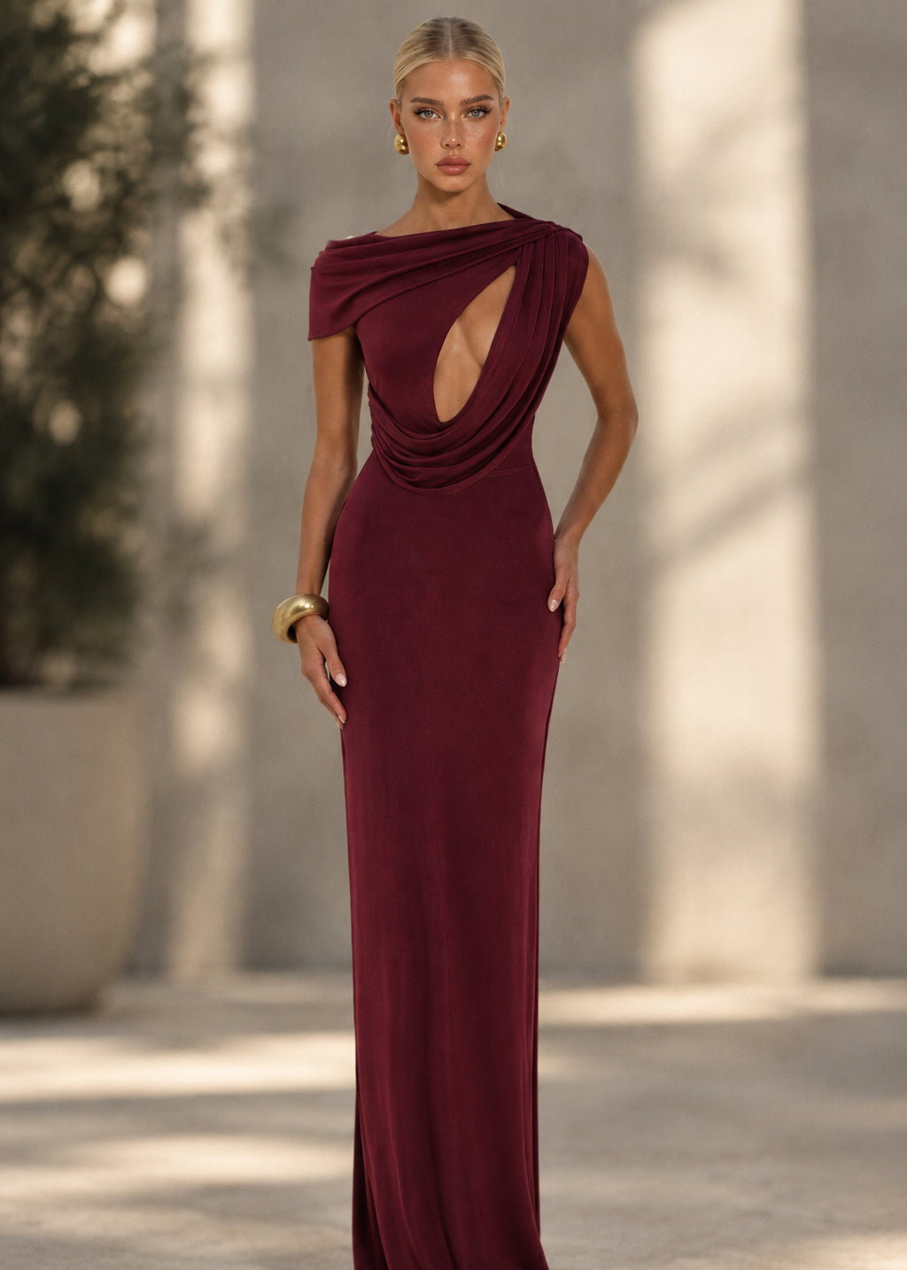 A woman modeling a floor-length, off-the-shoulder gown in deep burgundy with a dramatic draped neckline and a large cutout at the waist, posed against a plain light gray studio background.