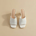 White open-toe mule high heels with a textured knit upper and chunky block heel, shown being worn and held by a hand against a light gray wood background.