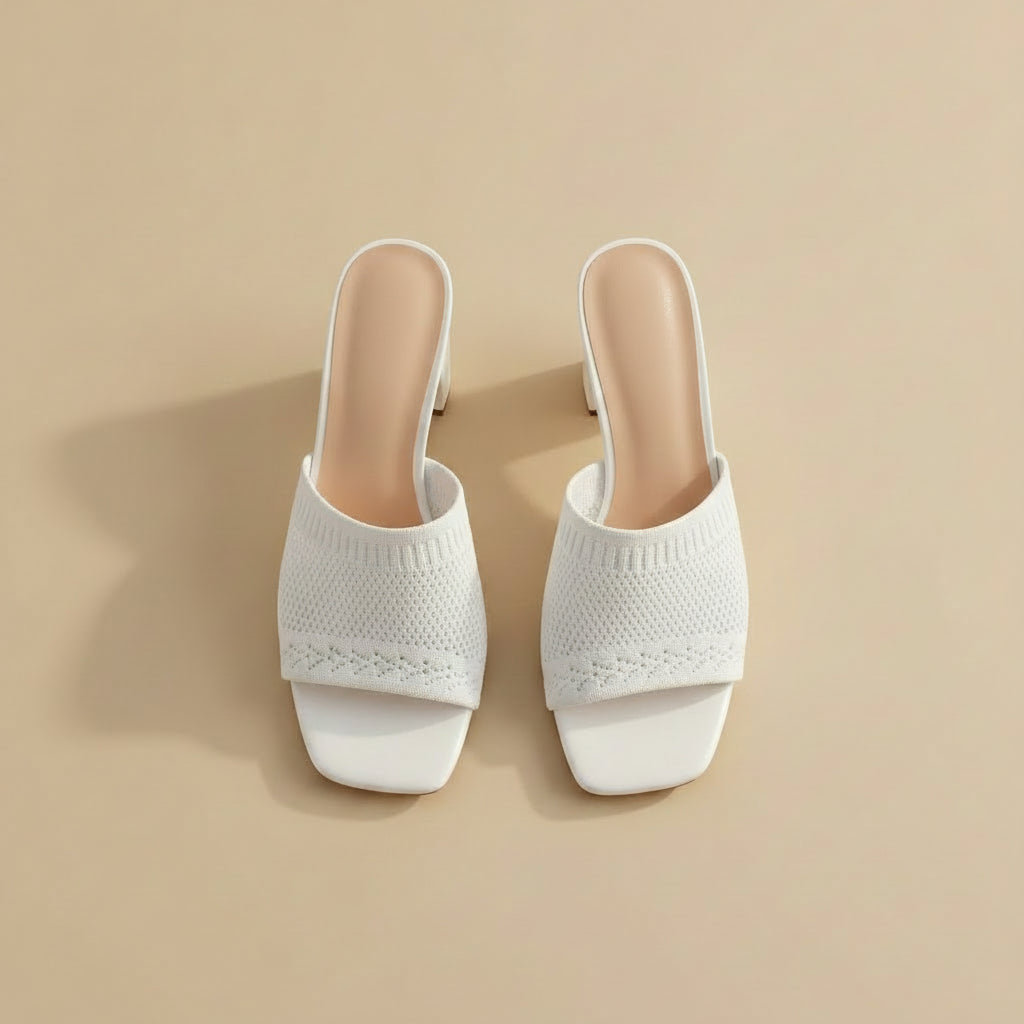 White open-toe mule high heels with a textured knit upper and chunky block heel, shown being worn and held by a hand against a light gray wood background.